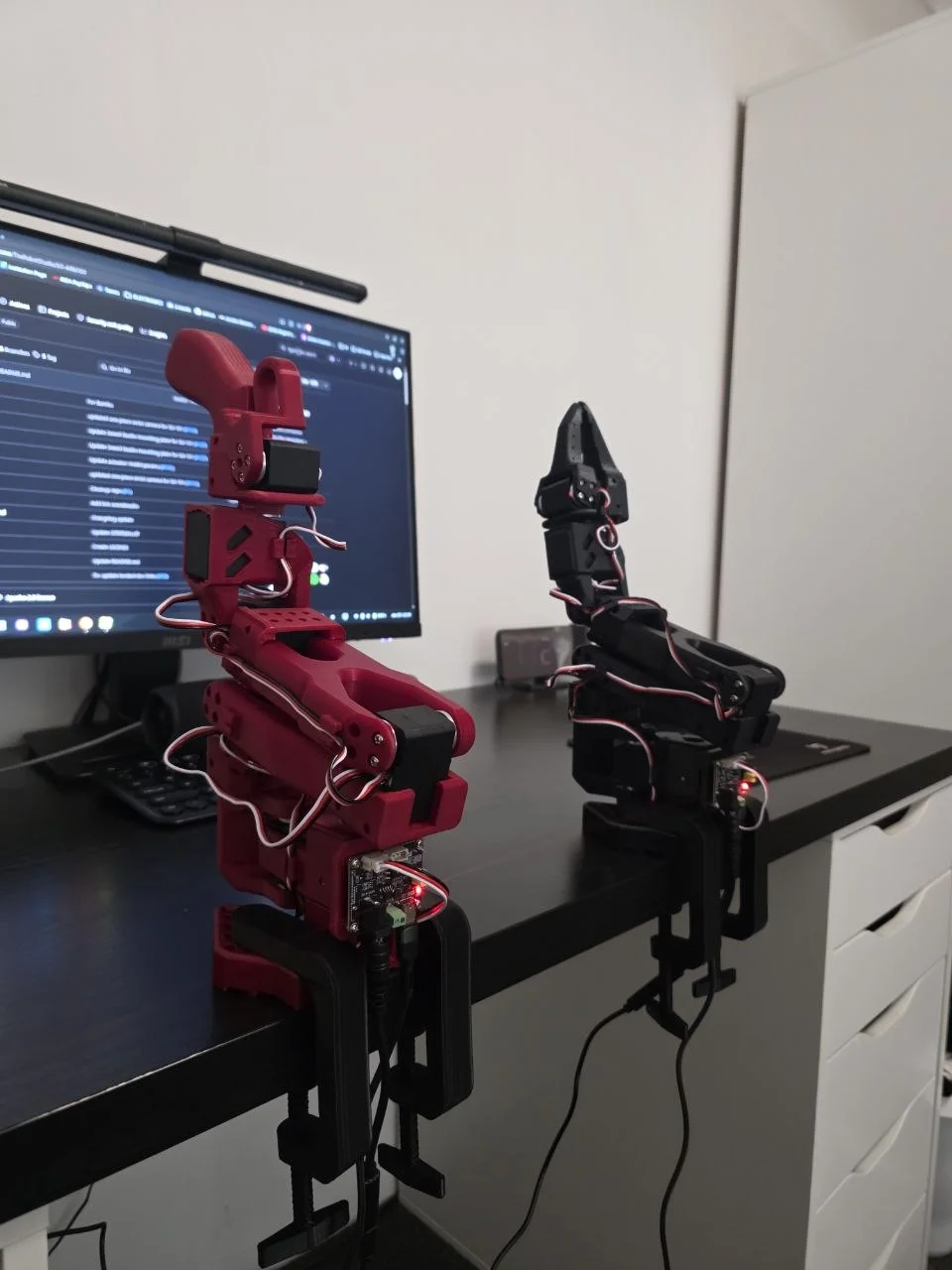 Leader (red) and follower (black) arms, desk-clamped, monitor with the SO-ARM100 repo in the background